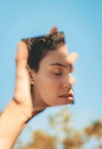 A serene woman holds a mirror with her reflection against a bright blue sky.