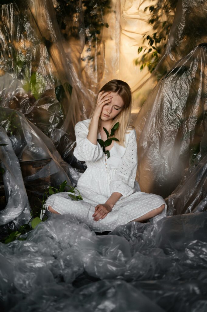 Thoughtful woman in white dress sitting amidst plastic and foliage indoors.
