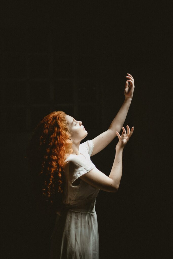 Elegant woman with red hair posing gracefully against a dark background, illuminated by soft light.