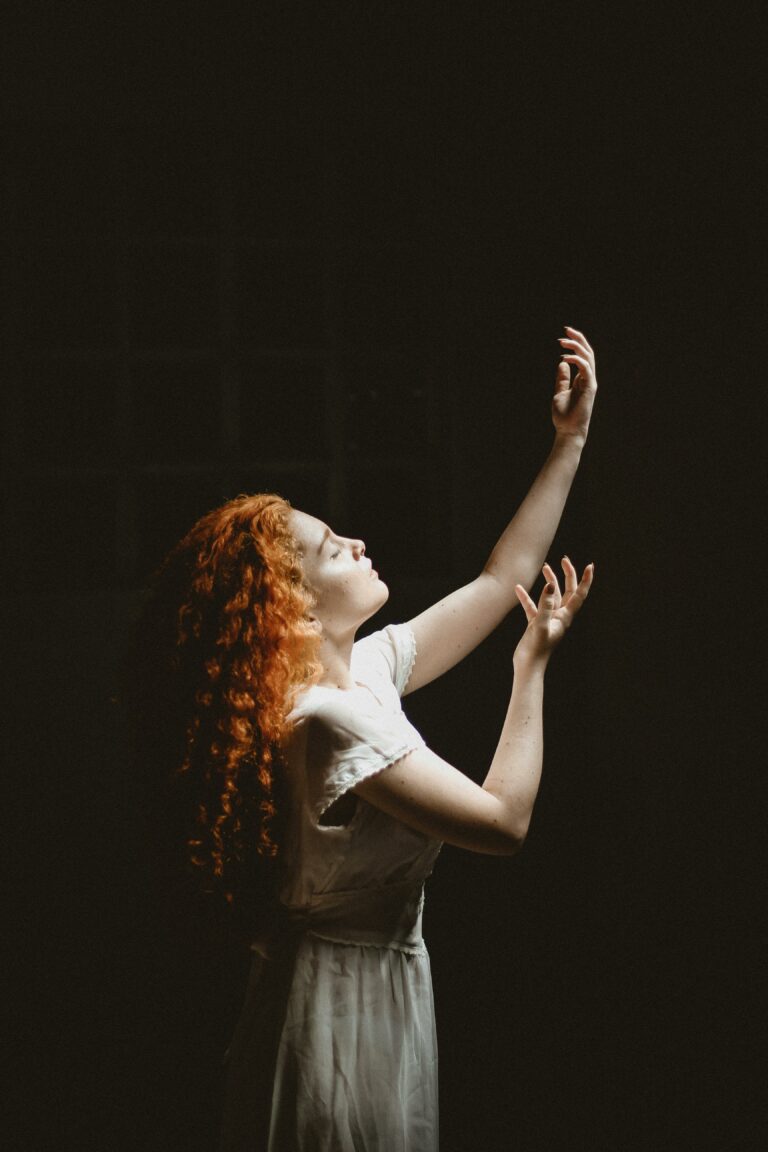 Elegant woman with red hair posing gracefully against a dark background, illuminated by soft light.