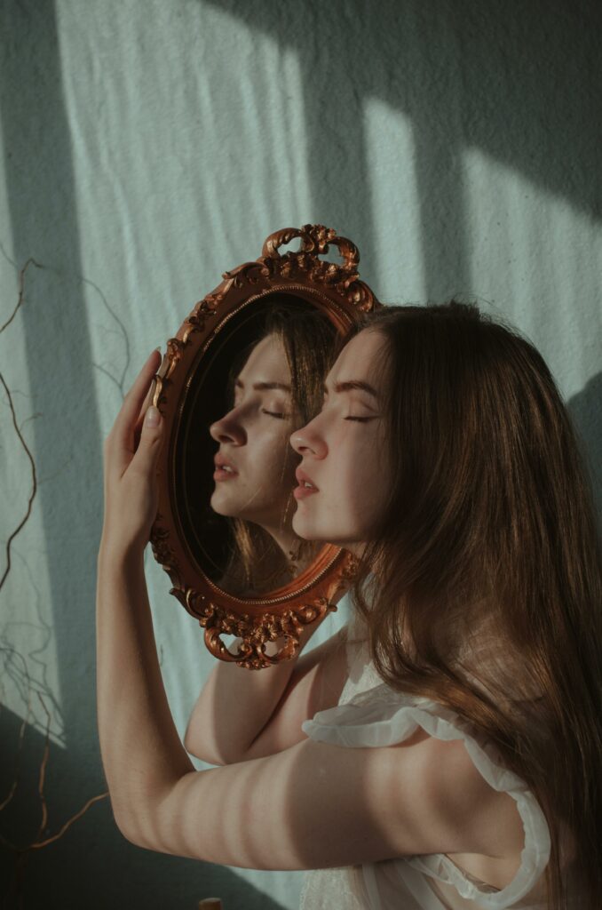 A young woman with eyes closed reflects in a vintage ornate mirror indoors.