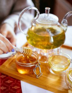 Elegant glass teapot with herbal tea, honey drizzle, and cups on a wooden tray for a perfect tea time setting.