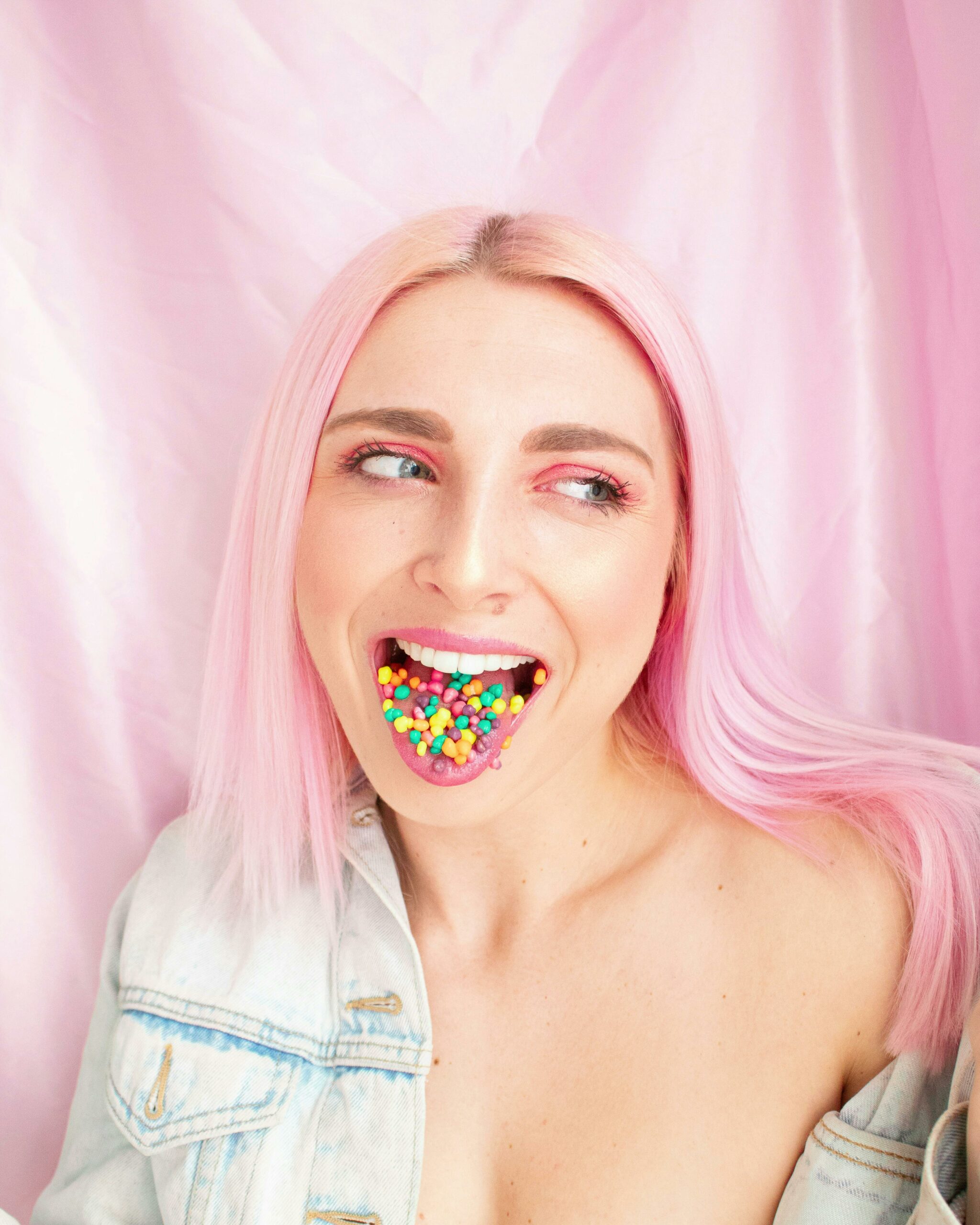 A woman with pink hair poses playfully with colorful candies in her mouth against a pink backdrop.