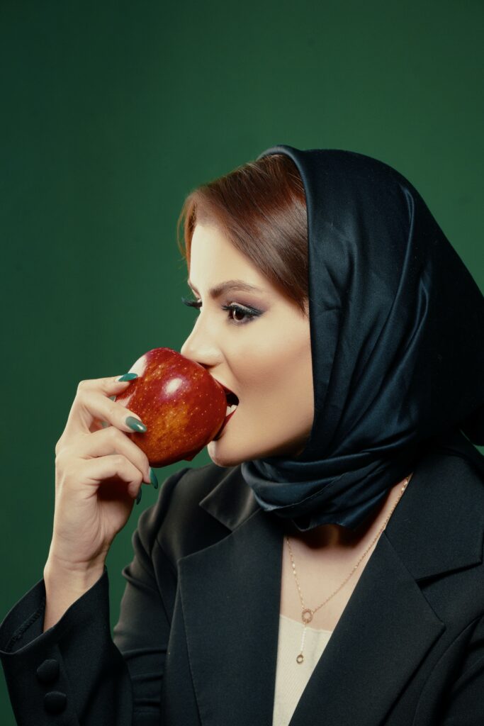 Woman in black headscarf and attire eats a red apple against a green background.