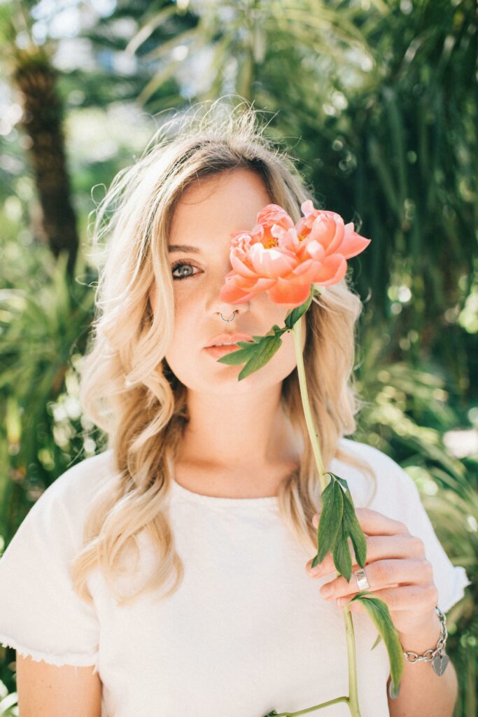 A blonde woman partially obscures her face with a pink flower in a lush outdoor setting.