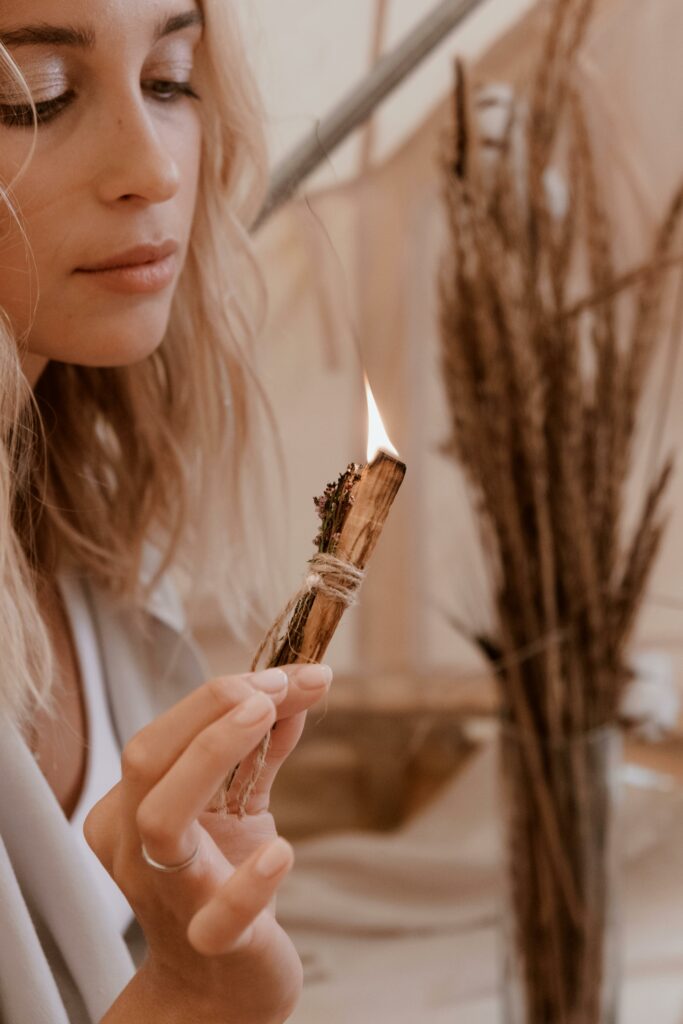 A serene woman engages in a purification ritual with a burning sage smudge stick indoors.