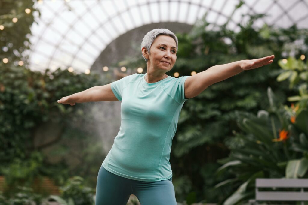 Elderly woman performing a yoga warrior pose in a lush green indoor botanical garden.