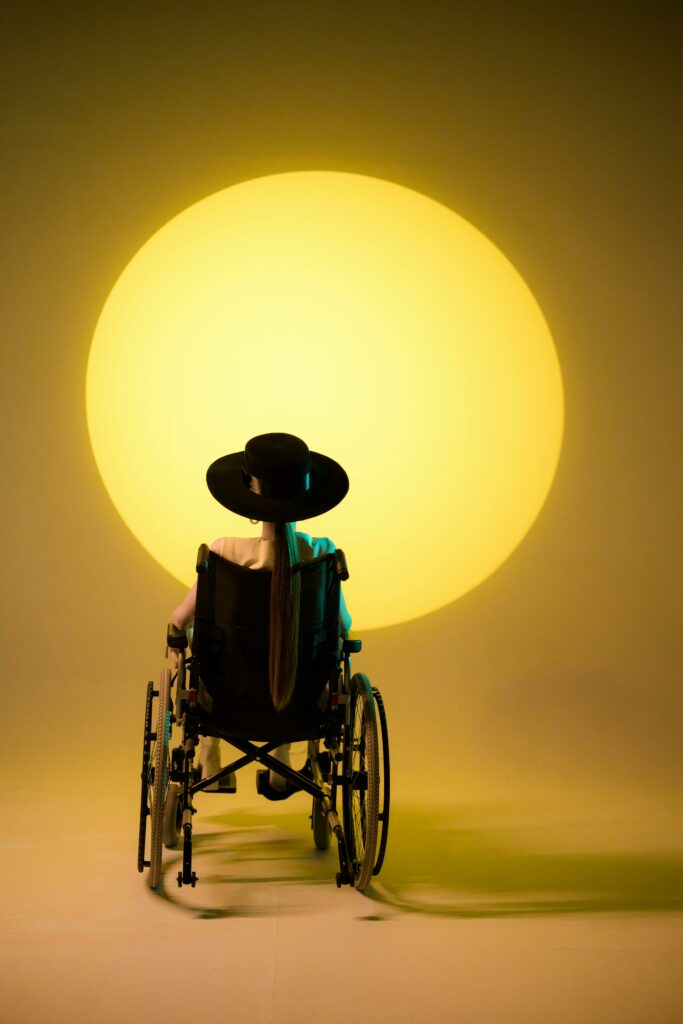 Woman in wheelchair wearing a hat, facing a large yellow spotlight, creates a contemplative mood.