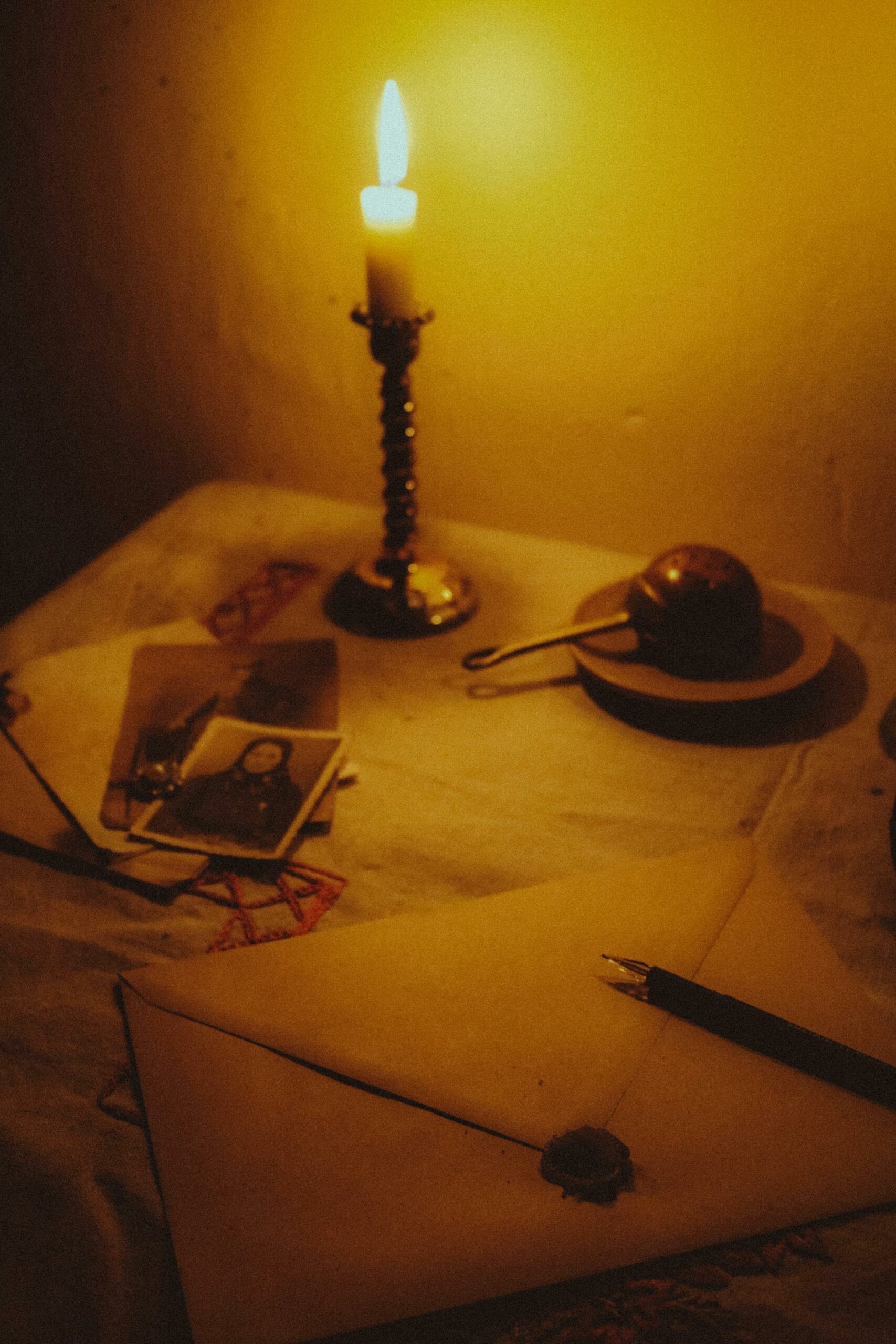 A vintage still life featuring a candlelit desk with a sealed envelope, fountain pen, and old photographs.