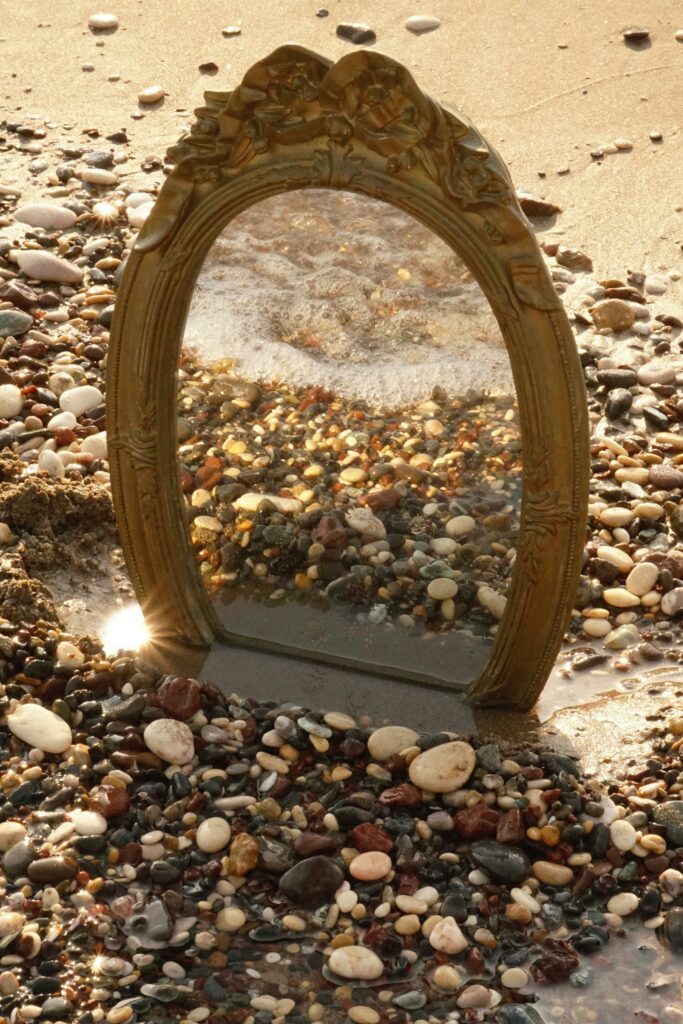 A vintage mirror reflects the vibrant pebble beach, creating an artistic and surreal shoreline scene.