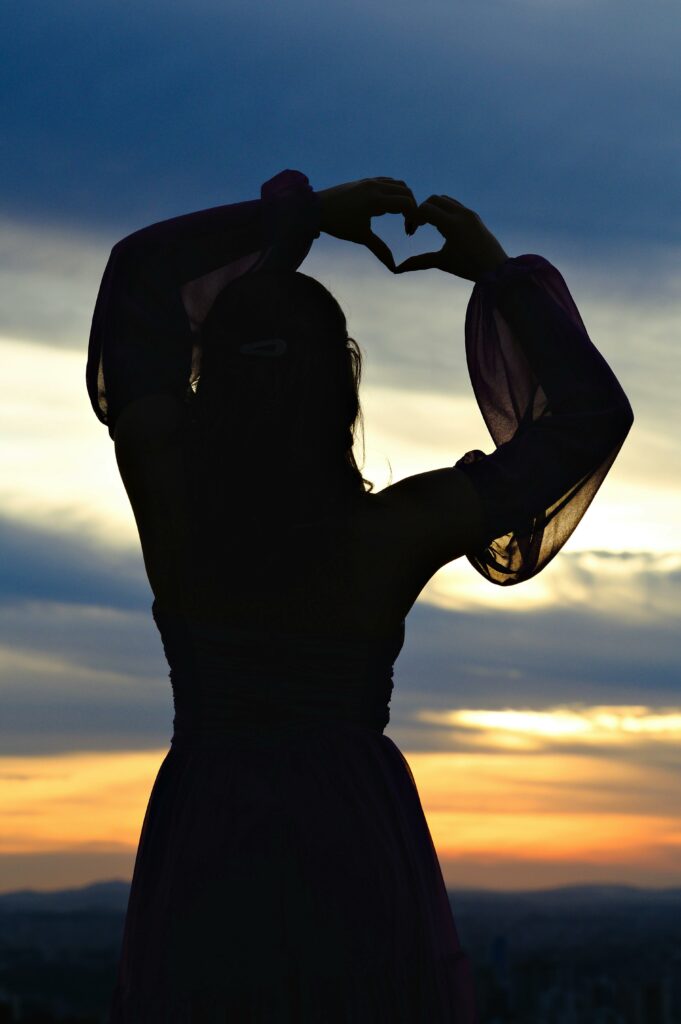 Silhouette of a woman forming a heart with hands against a stunning sunset backdrop.
