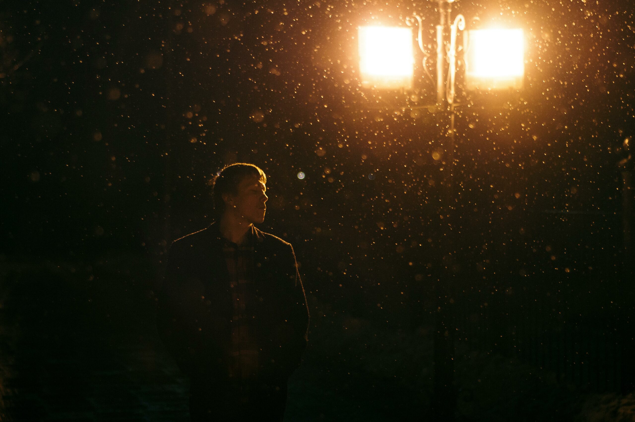 A man stands under a streetlight with snowflakes falling at night, creating a moody winter scene.