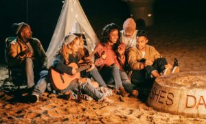 Group of friends enjoying music and socializing by a bonfire at night on the beach.