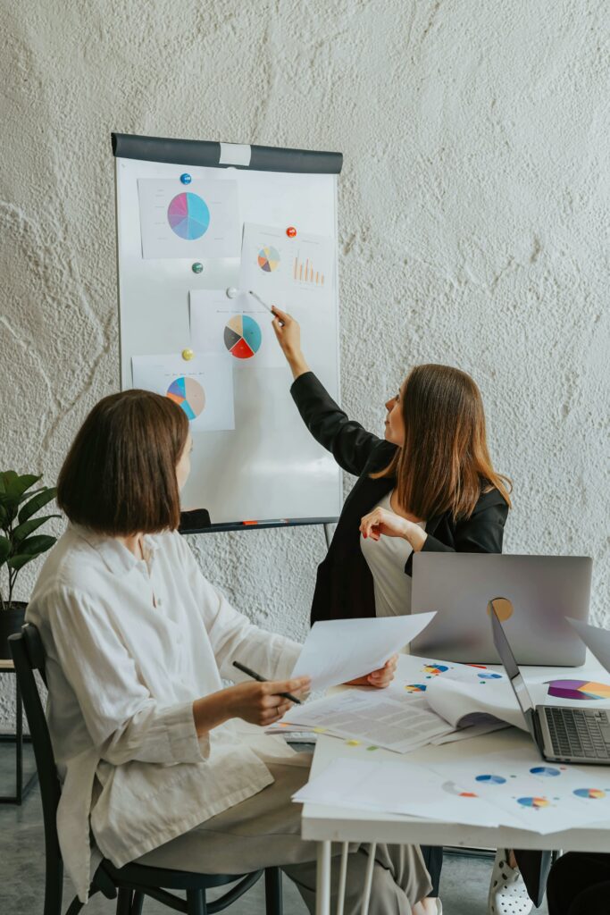 Two women collaborating in an office setting, reviewing charts and data on a flipchart.