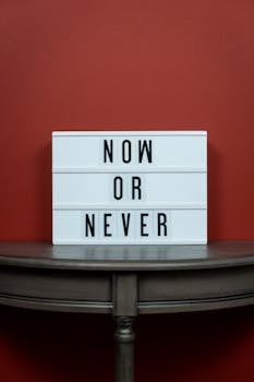 White letter board with 'Now or Never' text against a red wall, conveying urgency.