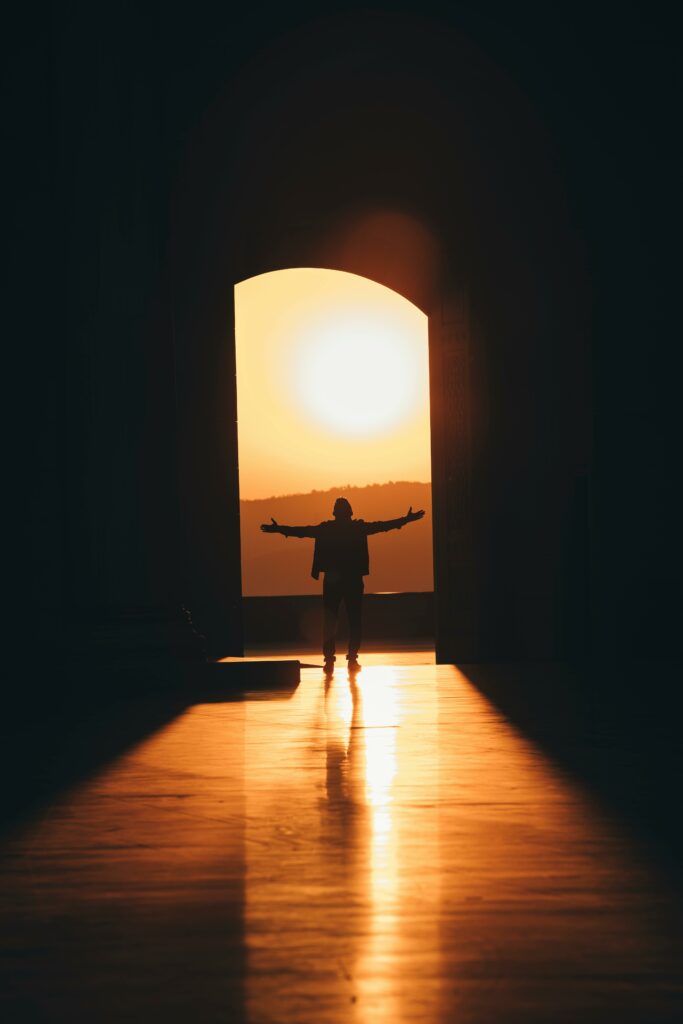 A person standing in a doorway with arms outstretched, silhouetted by a glowing sunset.