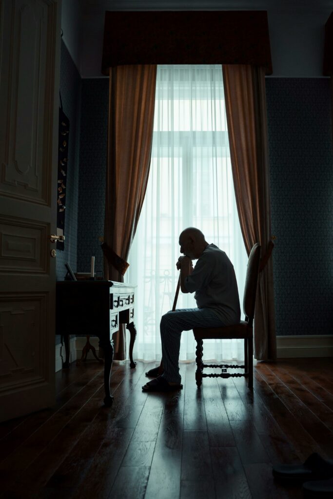 Elderly man sitting in thoughtful pose by a window in dimly lit room, exuding a serene atmosphere.