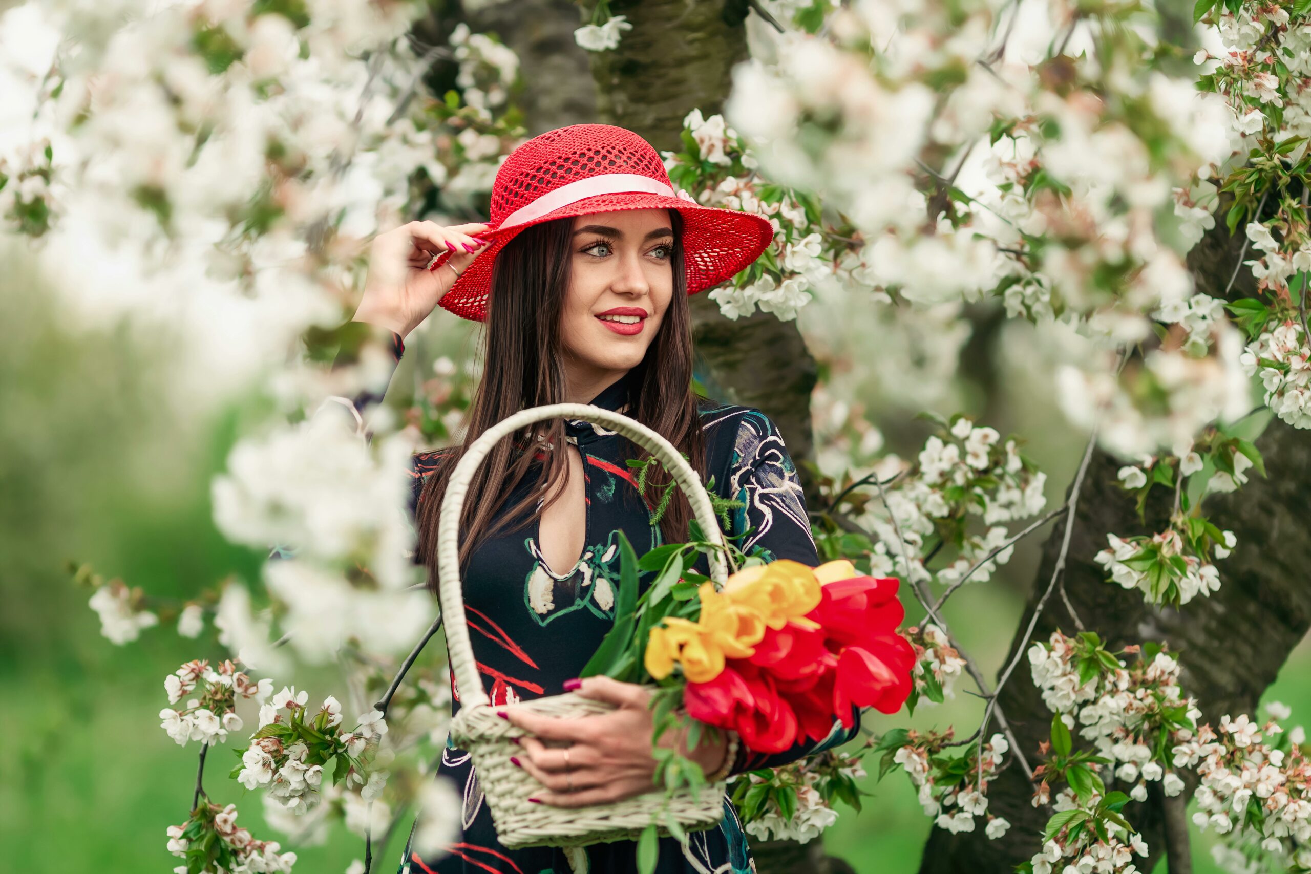 Woman with red hat standing among blossoming trees holding a basket of flowers.