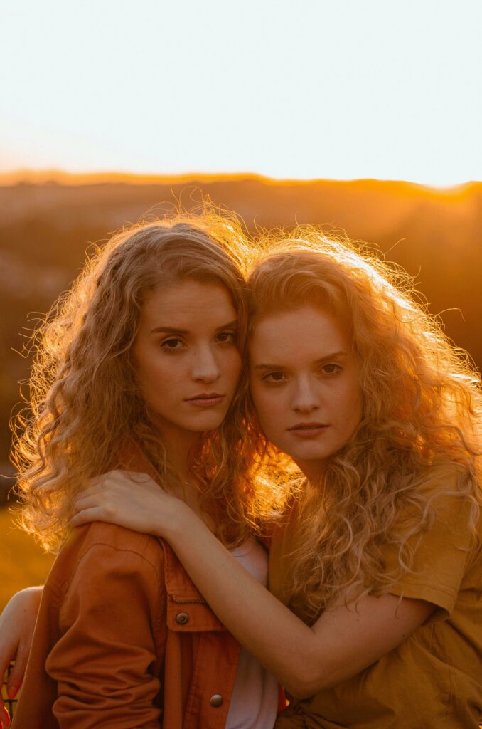 Two young women with curly hair embrace during a golden sunset.