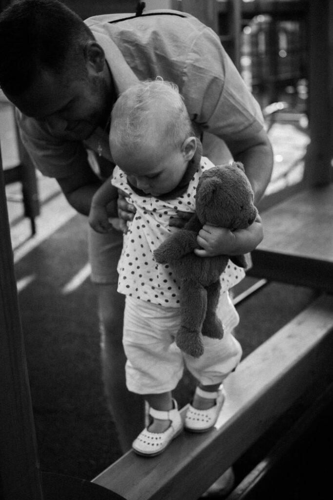 A father assists his baby in walking on a wooden beam, emphasizing balance.