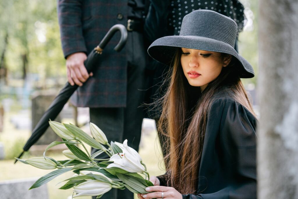 A woman in a black hat holds lilies, mourning at a cemetery.