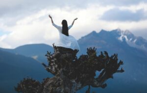 A woman in white stands on a tree stump with mountains in the background, expressing freedom and serenity.