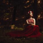 A young woman wearing an elegant red dress and flower crown sits in a serene forest setting, evoking fantasy and grace. pexels-photo-16564582-16564582