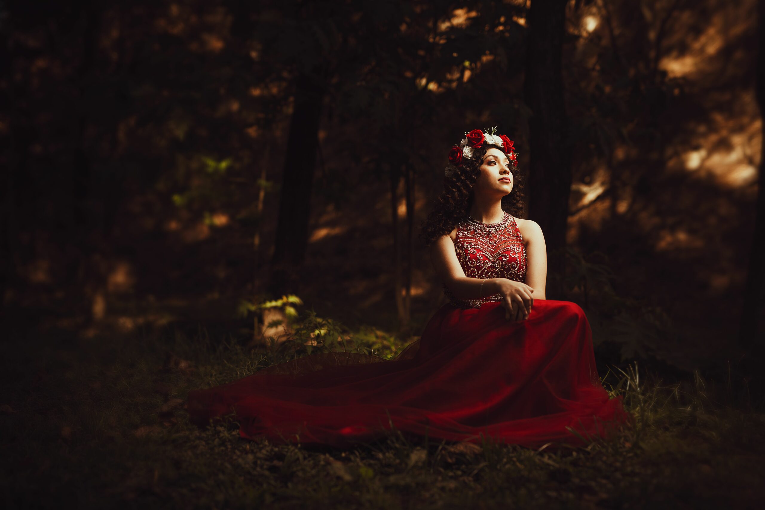 A young woman wearing an elegant red dress and flower crown sits in a serene forest setting, evoking fantasy and grace.