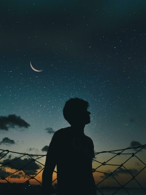 A lone silhouette stands beneath a crescent moon and starry sky, evoking a sense of solitude and wonder.