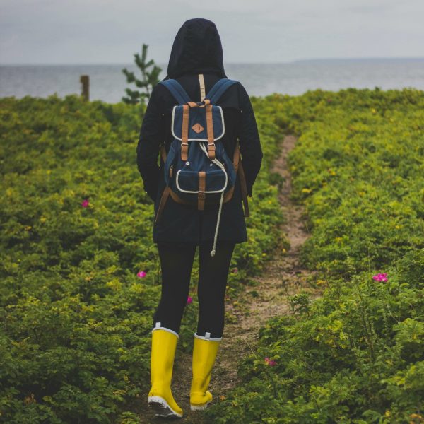 Person in yellow boots hiking through greenery towards the sea under cloudy skies.