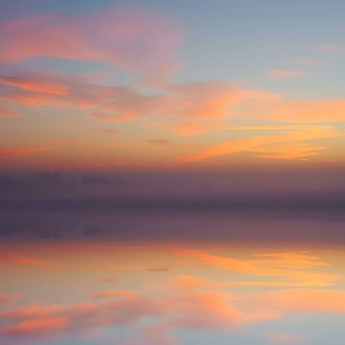Tranquil sunrise with colorful sky reflecting over calm waters and a lone tree.