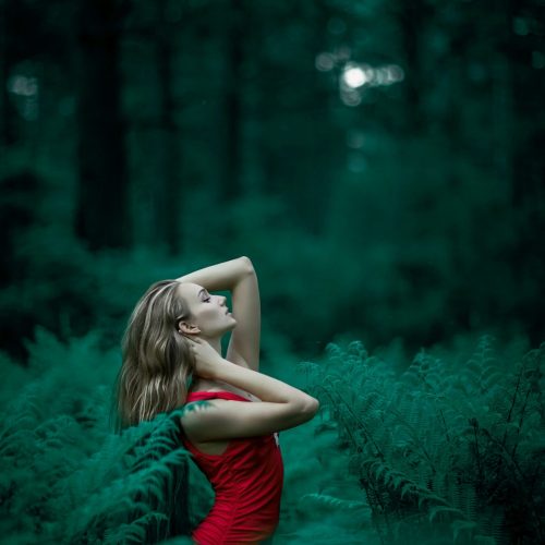 A woman poses in a vibrant red dress amidst lush green ferns in an enchanting forest setting.