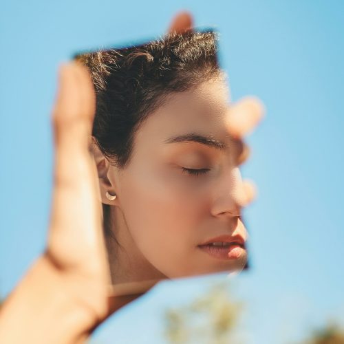 A serene woman holds a mirror with her reflection against a bright blue sky.