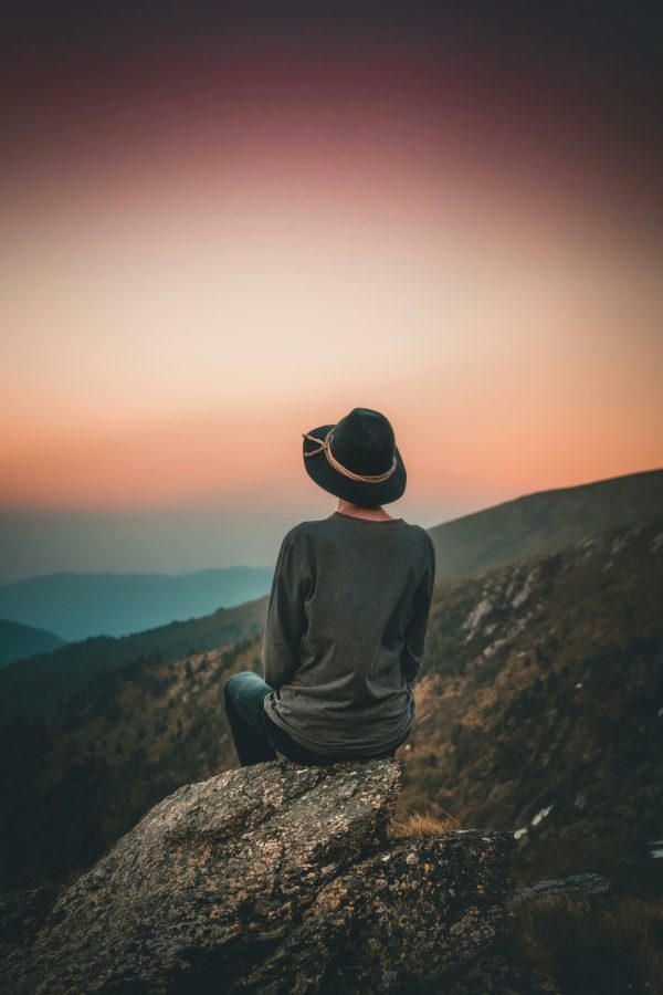 Person in hat sits on rock, gazing at sunset over mountains. Perfect for travel and nature themes.