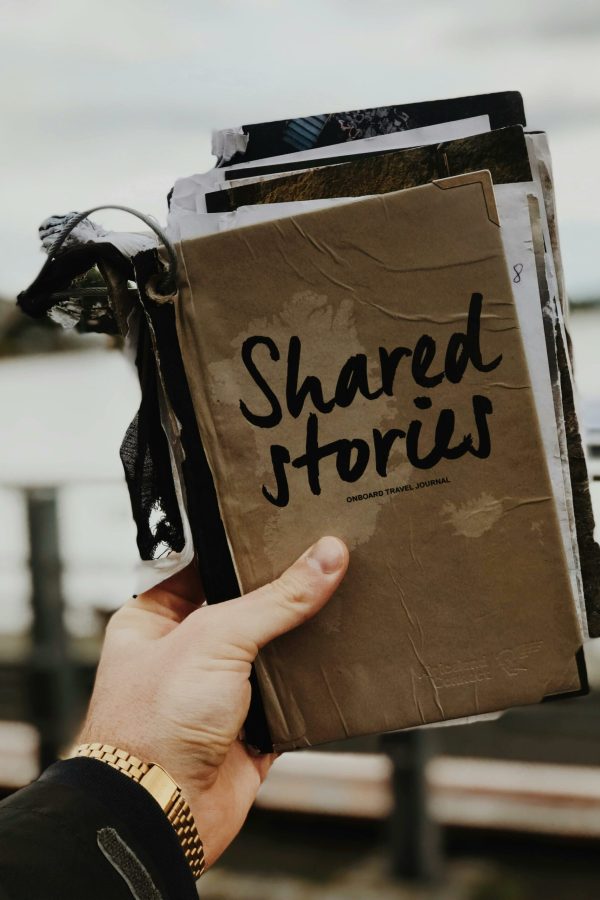 Hand holds a "Shared Stories" journal against a blurred outdoor backdrop, emphasizing creativity and personal storytelling.