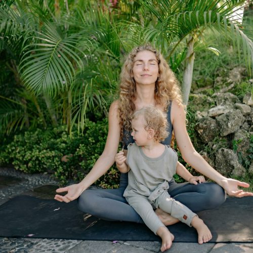Full body happy relaxed mother sitting with eyes closed in lotus pose with cute boy on laps while practicing yoga in tropical green park