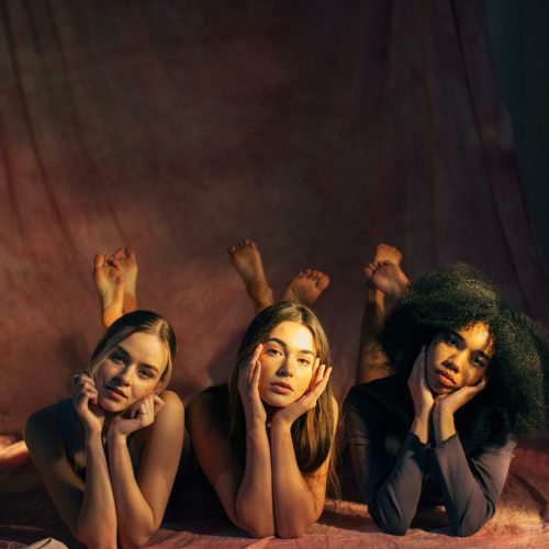 Three diverse women pose together on the floor in a studio, conveying empowerment and unity.