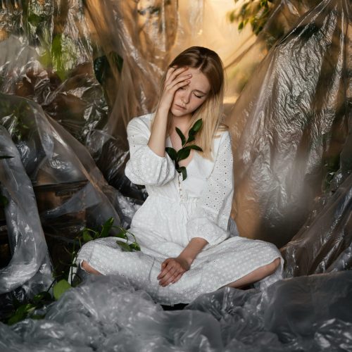 Thoughtful woman in white dress sitting amidst plastic and foliage indoors.